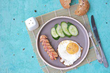 Kahvaltı, sosisli sahanda yumurta ve turkuaz beton arka planda kahverengi kil tabakta salatalık dilimleri. Çocuk kahvaltıları. Okul kahvaltı konsepti. Üst görünüm, alanı kopyala.
