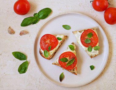 Açık sıcak sandviçler, mozarella peyniri, taze domates ve açık beton arka planda beyaz bir tabakta fesleğen. İtalyan yemeği. Üst görünüm, alanı kopyala.