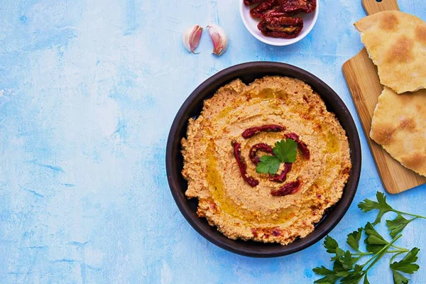 Hummus with sun-dried tomatoes in a brown clay bowl on a light blue concrete background. Served with tortillas, drizzled with olive oil when applying. Healthly food. Recipes chickpeas. Vegan food.