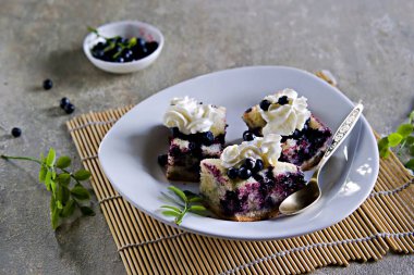Dilimlenmiş yaban mersinli kek, beyaz bir tabakta gri beton arka planda kremayla süslenmiş. Böğürtlenli kek. Mevsimlik tarifler. 