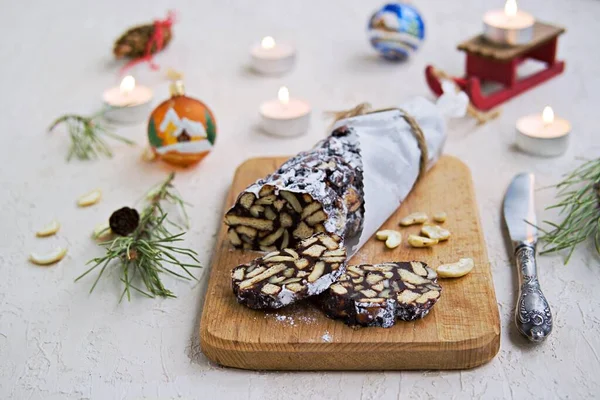 Dessert, chocolate salami with cookies and cashews in the New Year and Christmas style on a wooden board on a light concrete background. Desserts without baking.