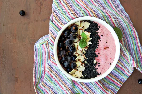 Smoothie bowl based on mashed berries, banana and natural yogurt in a white bowl. Decorated with frozen blackcurrant, sliced almonds and black sesame seeds. Healthy dessert. Top view, copy space.