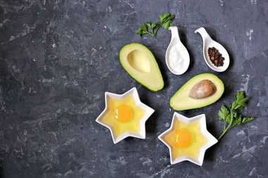 Koyu beton arka planda yumurtalı avokado pişirmek için gereken malzemeler: olgun avokado, ikiye bölünmüş yumurta, tuz, biber, maydanoz. Üst görünüm, alanı kopyala.