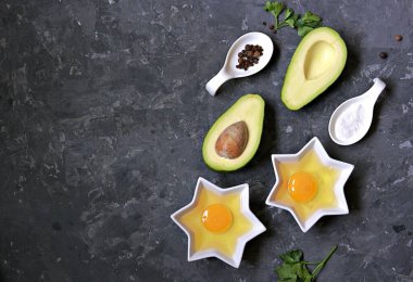 Koyu beton arka planda yumurtalı avokado pişirmek için gereken malzemeler: olgun avokado, ikiye bölünmüş yumurta, tuz, biber, maydanoz. Üst görünüm, alanı kopyala.