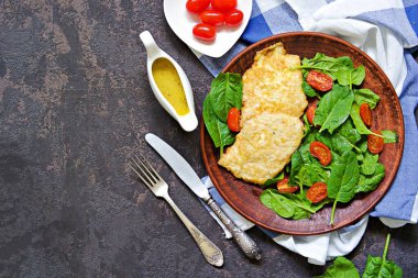 Koyu beton zeminde kahverengi bir tabakta hindi göğsü doğrar. Yanında taze ıspanak ve vişneli domates salatası. Üst görünüm, alanı kopyala.