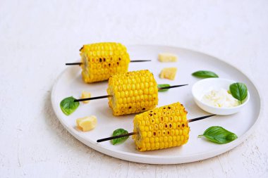 Beyaz seramik tepside ızgara mısır şişi. Tuzlu tereyağı ve rendelenmiş parmesan peyniri ile servis edilir. Sebze tabakları. Mısırları topla..