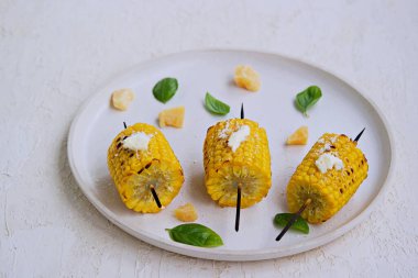 Beyaz seramik tepside ızgara mısır şişi. Tuzlu tereyağı ve rendelenmiş parmesan peyniri ile servis edilir. Sebze tabakları. Mısırları topla..