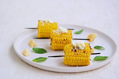 Beyaz seramik tepside ızgara mısır şişi. Tuzlu tereyağı ve rendelenmiş parmesan peyniri ile servis edilir. Sebze tabakları. Mısırları topla..