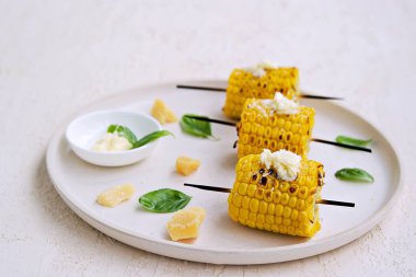 Beyaz seramik tepside ızgara mısır şişi. Tuzlu tereyağı ve rendelenmiş parmesan peyniri ile servis edilir. Sebze tabakları. Mısırları topla..