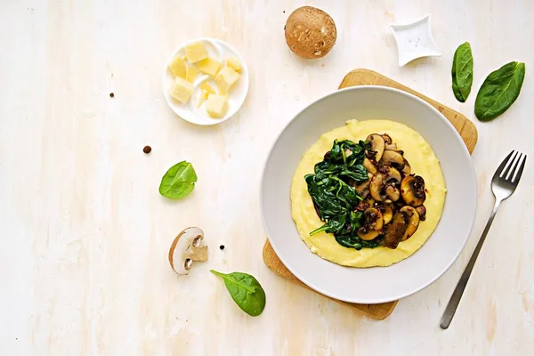 A portion of polenta or corn grits with fried mushrooms and stewed spinach in a gray plate on a light concrete background. Polenta or corn grits recipes. Italian Cuisine.