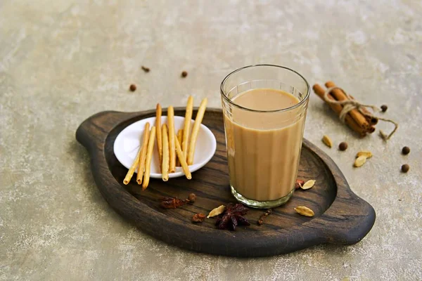 Hot drink, masala tea with milk in a faceted glass on a wooden brown tray on a gray concrete background. Served with salty crackers. Indian food, drinks. Spicy tea recipes.