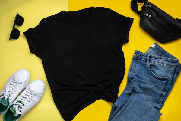 Sarı arka planda siyah tişört modeli. Hipster elbiseli düz yatak. Mavi kot, siyah güneş gözlüğü, beyaz spor ayakkabı ve omuz çantası.