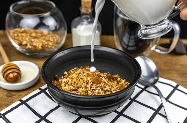 Выливание молока в черную миску домашней мюсли с орехами, овсяными хлопьями, изюмом. Концепция здорового питания. Фитнес завтрак
