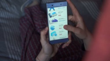 Akıllı telefonu tutan, sayfaları karıştıran bir elin yakın çekimi. Tuvalet kağıdı, el dezenfektanı, dezenfektan mendil, yüz maskesi, covid-19 nedeniyle lateks eldiven gibi ihtiyaçlar için online alışveriş.