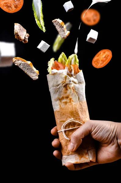 Shawarma tavuğu. İçinde malzemeler olan bir elde yuvarlan. Siyah arka planda havada uçuşuyor.