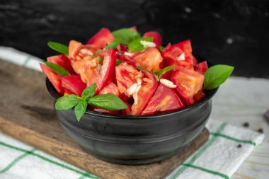 Olgun domates dilimleri ve yeşil fesleğen salatası. Tahta kesme tahtasının üzerinde siyah bir kasede.