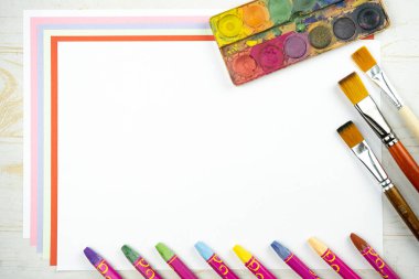 Düz zemin üzerinde beyaz kağıt çarşaf, palet suluboya boya boya, fırça, renkli tebeşir boya kalemleri, üst görünüm