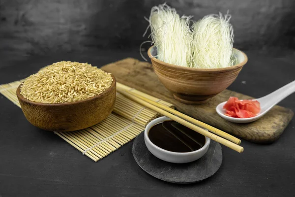 Tahta bir kasede kahverengi pirinç, bambu hasırlı Asya mutfağı arka planı, yemek çubukları, soya sosu ve siyah arka planda cam pirinç eriştesi.
