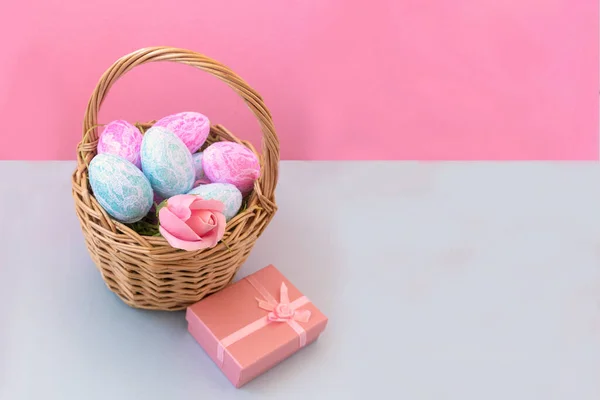 Basket with Easter eggs on a delicate pink background, next to a pink box with a gift. Easter holiday concept