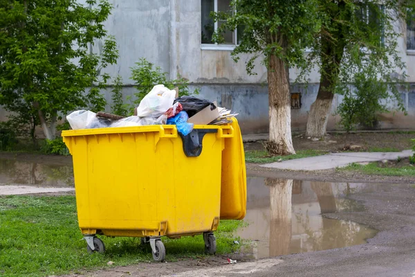 Trash bin, garbage container full of waste. Yellow trash can near the apartment building. Environmental pollution concept, bad ecology.