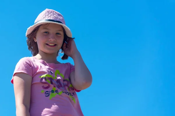 戴着粉色帽子的漂亮小女孩微笑着看着摄像机 在明亮的蓝天的背景下 快乐的女孩 低角度射击 复制空间 — 图库照片