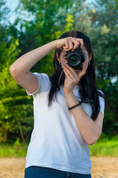 A beautiful brunette holds a SLR camera in her hands and looks into the lens. Taking pictures on a summer day in the park.
