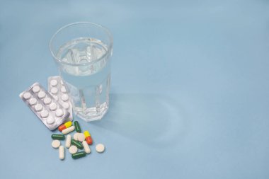 Beyaz haplı kabarcık, şeffaf bir bardak su ve çeşitli haplar ve kapsüller mavi arka plana saçılmış. Uzayı kopyala, seçici odaklanma. Eczacılık konsepti, sağlık hizmetleri.