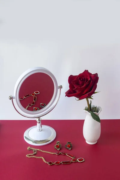Ruby gemstone jewelry set with reflection in vintage white mirror. Red rose in white vase on the table, beautiful accessories. Copy space, soft focus.