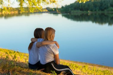 Sonbaharda nehrin yanındaki çimlerin üzerinde iki genç kadın oturuyor. Kız arkadaşlar sarılıyor, arkadan bakıyor..