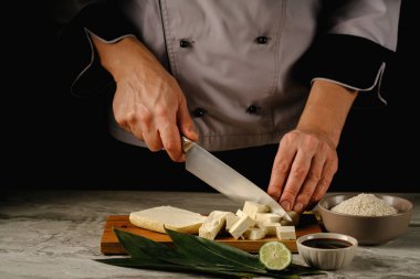 Bambu yaprakları üzerinde taze tofu parçaları, ahşap bir tahta üzerinde zencefil ve limon. Siyah bir arka planda. Tekstür için alan. Aşçının ellerinde tofu.