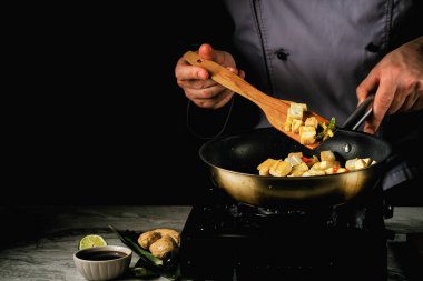 Bambu yapraklarının yanında taze tofu, soya sosu, zencefil ve limon. Aşçı tavada tofu keser. Tahta kaşıkla karıştırır.