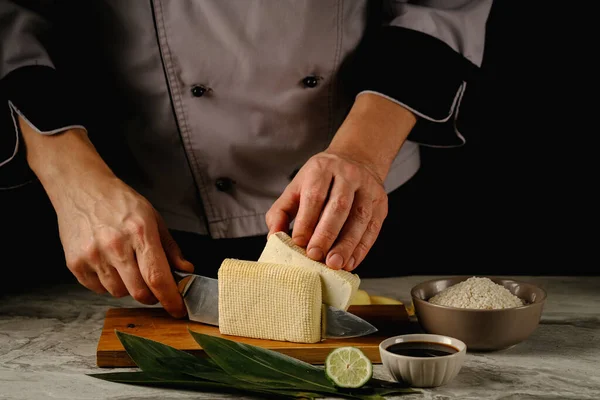 Pezzi di tofu fresco su foglie di bambù, su una tavola di legno zenzero e lime.On uno sfondo nero.Spazio per text.tofu nelle mani del cuoco — Foto Stock