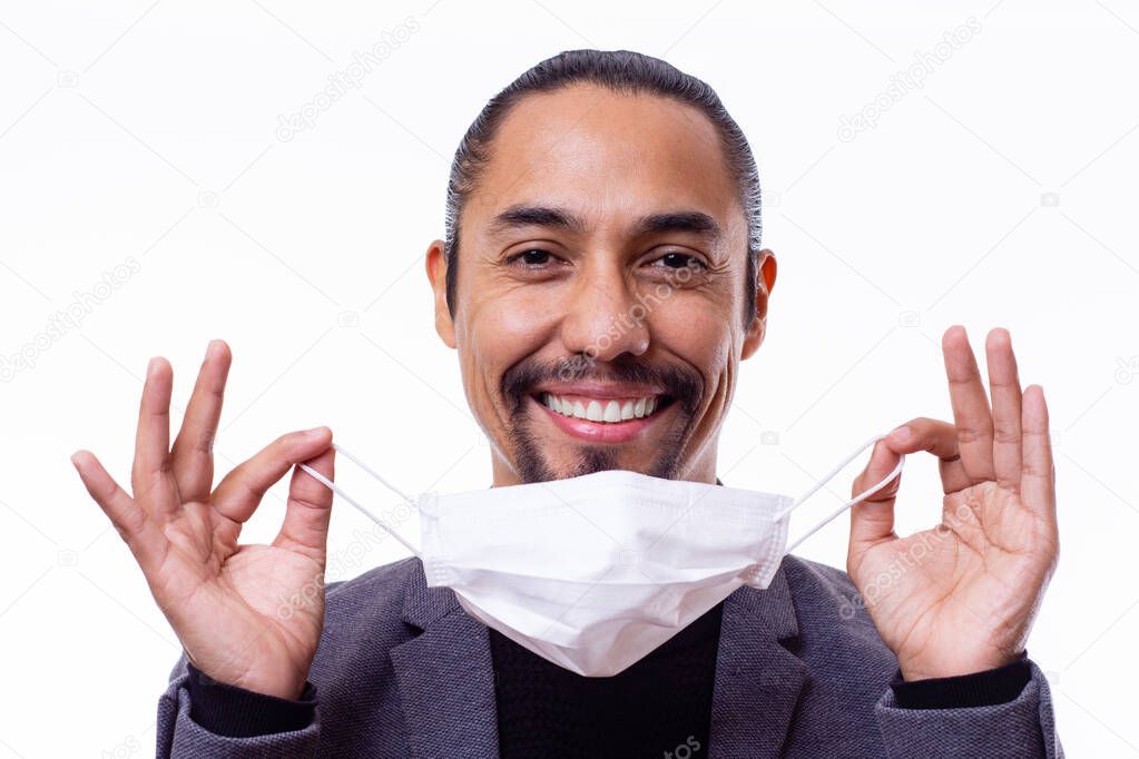 Close up of a latin man smiling happily while holding a medical mask with both hands on white background.