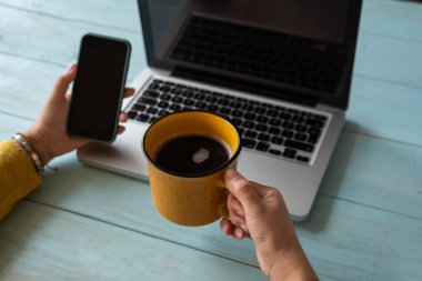 Akıllı telefon ve ahşap bir masanın üzerinde bir fincan kahveyle kadın eli seçici bir odak noktası. Ev konseptinde çalış.