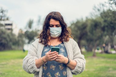 Kadın yüzünü koruyucu bir maskeyle örtüyor. Telefonu olan kadın sahada akıllı telefonunu kullanıyor. Coronavirus konsepti.