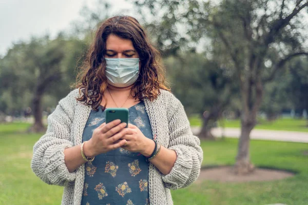 Kadın yüzünü koruyucu bir maskeyle örtüyor. Telefonu olan kadın sahada akıllı telefonunu kullanıyor. Coronavirus konsepti.