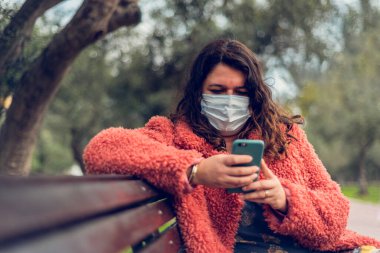 Parkta oturan bir kadın, akıllı telefonuyla yazarken tıbbi bir maskeyle kendini koruyor..