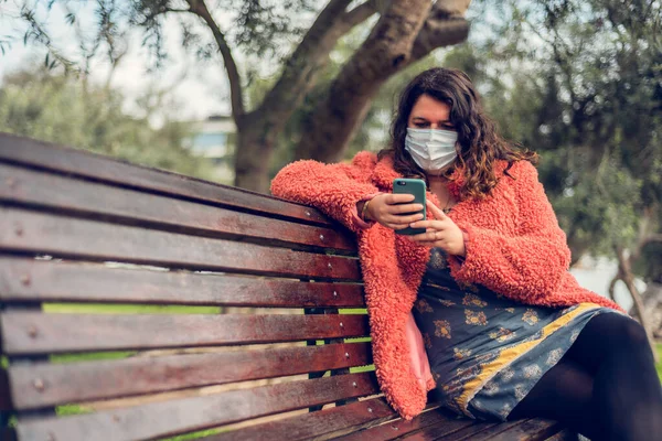 Tıbbi maskeli güçlü bir kadın akıllı telefonuna dışarıdaki parkta bir bankta oturuyor. Yeni normal konsept. Boşluğu kopyala