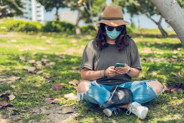 Kadın dışarıda otururken akıllı telefonuna bakıyor ve kendisini tıbbi bir maskeyle koruyor. Yeni normal