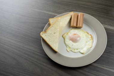 Kahvaltı beyaz tabakta, sağ alt köşede, ahşap zeminde.