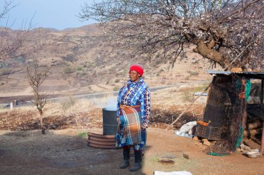 Ramabanta, Lesotho - 20 Eylül 2017: Geleneksel battaniye giysili yetişkin Afrikalı kadın köy avlusunda milli içeceği metal fıçıda demliyor, yaşlı kadın yemek hazırlıyor, yani insan hayatı.