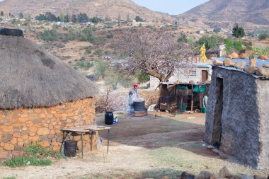 Ramabanta, Lesotho - 20 Eylül 2017: Geleneksel battaniye giysili yetişkin Afrikalı kadın köy avlusunda milli içeceği metal fıçıda demliyor, yaşlı kadın yemek hazırlıyor, yani insan hayatı.