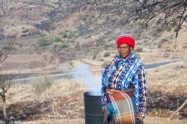 Ramabanta, Lesotho - 20 Eylül 2017: Geleneksel battaniye giysili yetişkin Afrikalı kadın köy avlusunda milli içeceği metal fıçıda demliyor, yaşlı kadın yemek hazırlıyor, yani insan hayatı.