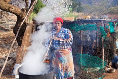Ramabanta, Lesotho - 20 Eylül 2017: Geleneksel battaniye giysili yetişkin Afrikalı kadın köy avlusunda milli içeceği metal fıçıda demliyor, yaşlı kadın yemek hazırlıyor, yani insan hayatı.