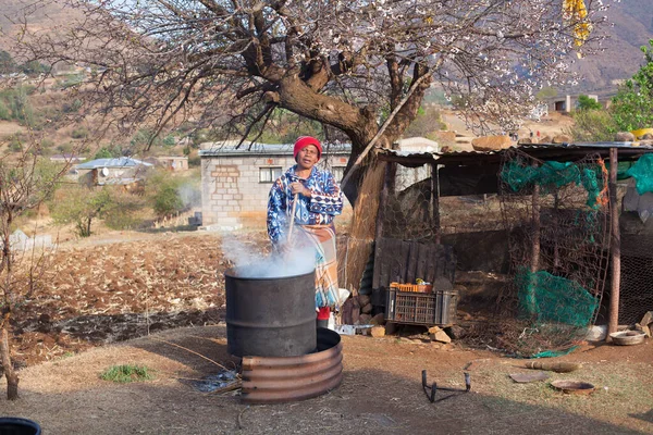 Ramabanta, Lesotho - 20 Eylül 2017: Geleneksel battaniye giysili yetişkin Afrikalı kadın köy avlusunda milli içeceği metal fıçıda demliyor, yaşlı kadın yemek hazırlıyor, yani insan hayatı.