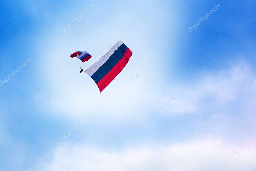 Russian paratrooper jumps with a parachute painted in the colors of the flag of Russia with the Russian flag on clear blue sky and white clouds background. Russian armed forces, landing troops