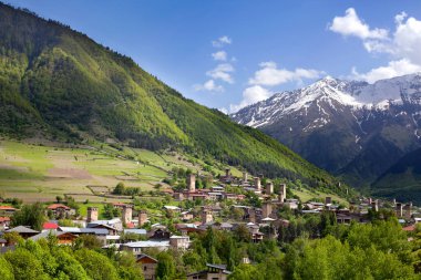 Gürcistan 'daki Ushguli köyü, Svaneti bölgesi, yeşil bir tepede antik kuleler Kafkas dağları, kardaki dağ zirveleri, mavi bulutlu arka plan