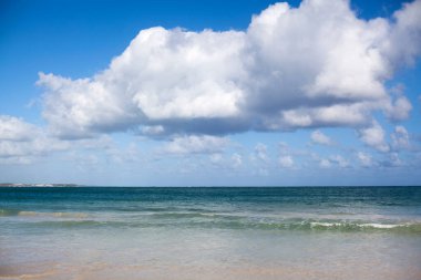 Kumsalda bir kadın, mavi bir denizde, mavi bir gökyüzünde ve büyük beyaz bir bulutun arkasında uzanıyor.