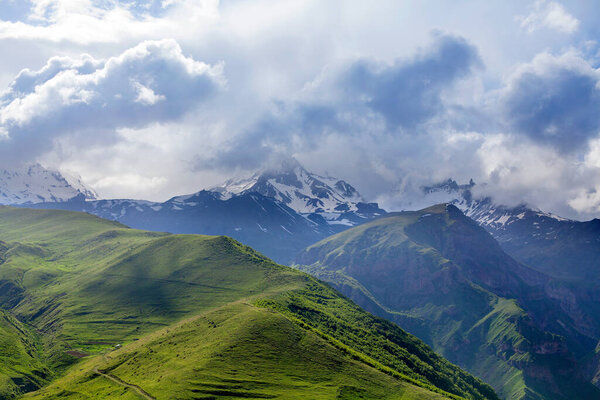 Пейзажи горных вершин в снегу и зеленые холмы, глубокое голубое небо и огромные белые облака фона, Кавказские горы, Казбек горы, Грузия
