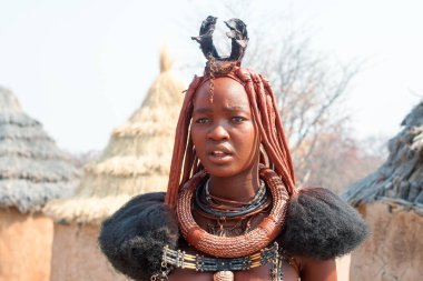 Otjikandero Himba Köyü, Namibya - 8 Eylül 2017: Himba Çamurlu kil evlerde geleneksel saç stiline, yüzüklere ve kolyeye sahip güzel Himba kadını